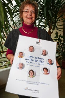 Barbara Burges von der Kontakt- und Informationsstelle für Selbsthilfegruppen des Kreises Soest präsentiert das Plakat „Wir leben Selbsthilfe im Kreis Soest! – Wir und viele andere“. Zum 15-jährigen Bestehen des Arbeitskreises Selbsthilfe ließen sich sieben Mitglieder ablichten (Foto: Judith Wedderwille/Kreis Soest).