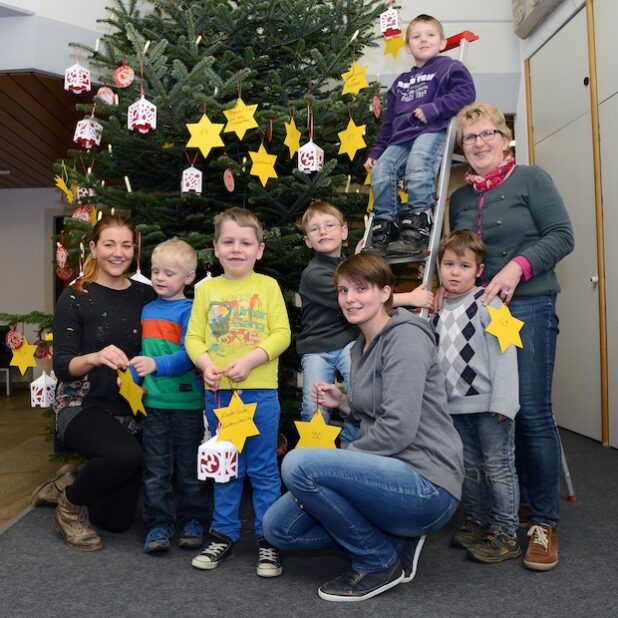 Mit gelben Sternen und weißen Laternen schmückten sechs Kinder der Heilpädagogischen Kindertageseinrichtung „Zwergenland“ den Weihnachtsbaum im Foyer des Kreishauses. Beim Aufhängen halfen Heilpädagogin Birgit Heinrich-Kleinschmidt (r.), Larissa Krauße (Freiwilliges soziales Jahr) und Erzieherin Corinna Oberley (knieend) - Foto: Thomas Weinstock/Kreis Soest.