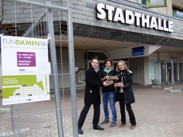 HSK-Wirtschaftsförderer Michael Bison (l.), Julia Reuther (M.) und Carmen Kieserling (r.) präsentieren das Plakat der „Fundamenta“ vor der Mescheder Stadthalle (Foto: WFG HSK).